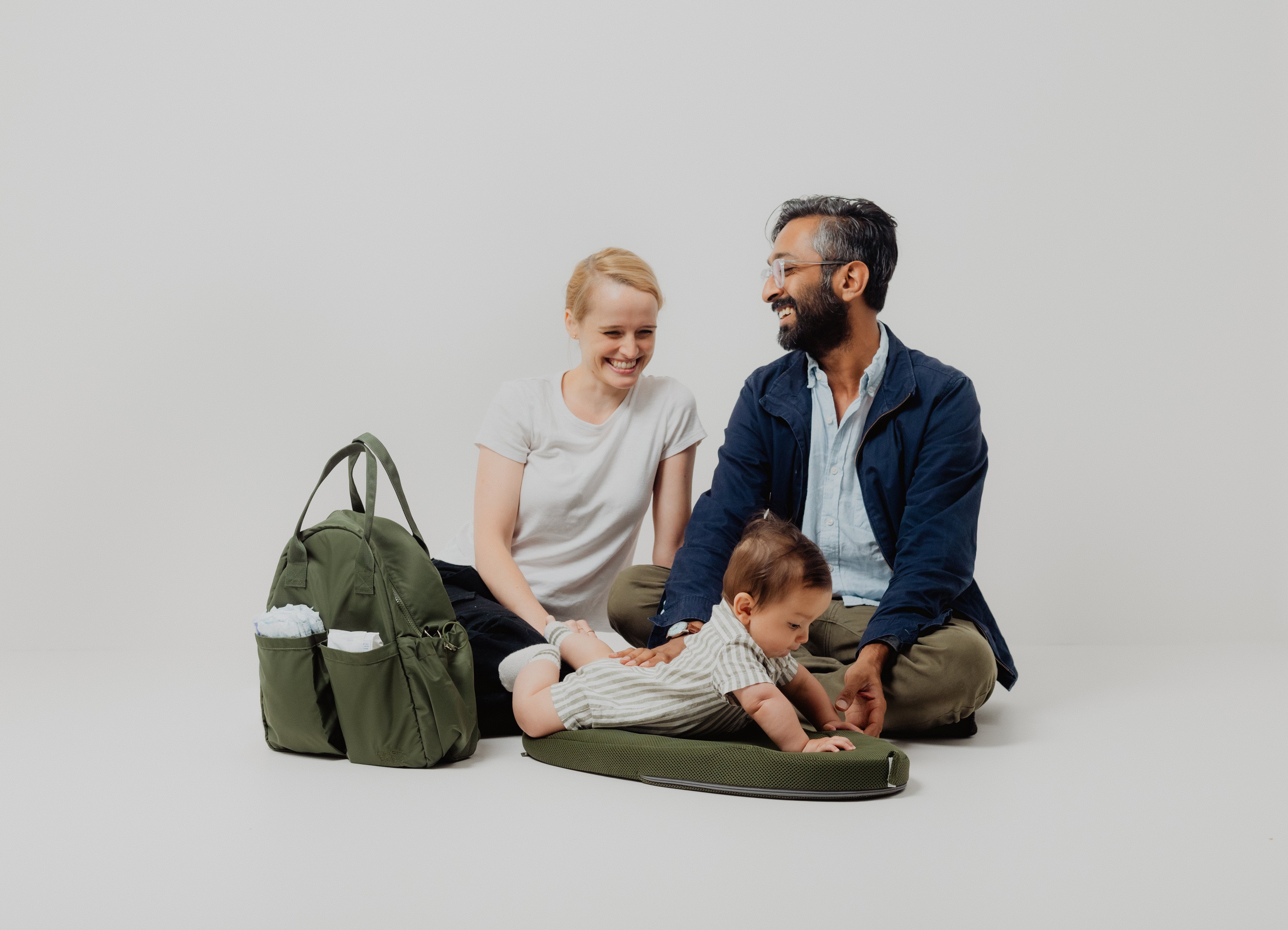 Family with a baby sitting on a green cushion with a green bag next to them against a white background
